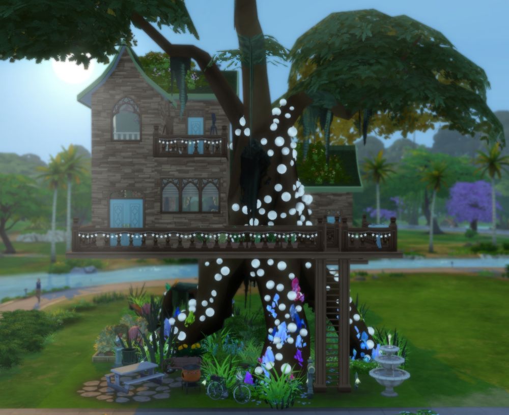 The front of the treehous in the morning. Round lights and crystals cover the tree trunk with plants underneath. A picnic table is under the tree with a grill and a garden is just visible next to the tree.