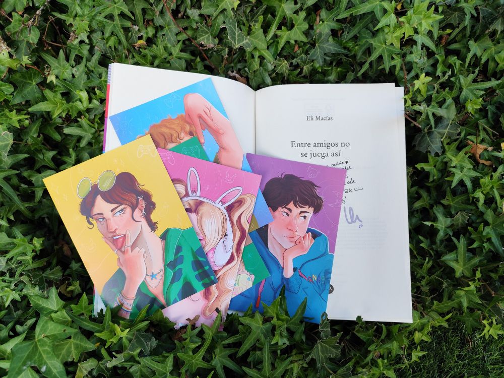 libro "entre amigos no se juega asi" de eli macias abierto por la portadilla, se ven unas postales de los personajes dibujadas por @ponypokez desplegadas encima además de hojas verdes en el fondo 