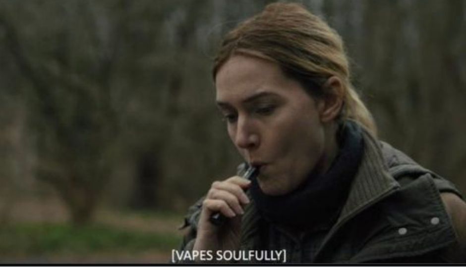 Kate Winslet in Mare of Easttown sucks on her vape while seated outside wearing a turtleneck and a green parka. The closed caption reads [VAPES SOULFULLY]