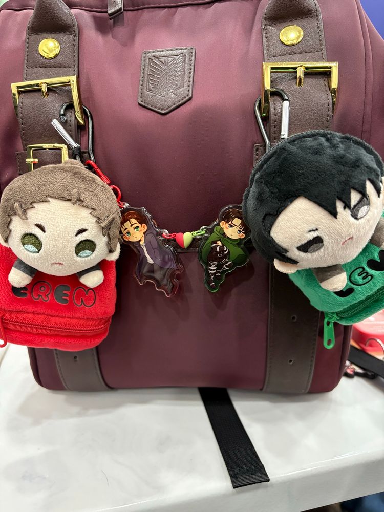 TJ’s backpack, an Eren-themed burgundy SUPER GROUPIES Attack on Titan backpack featuring Eren and Levi plushies dangling from it, as well as a pair of Eren and Levi keychains connected together by half red and green heart magnets.