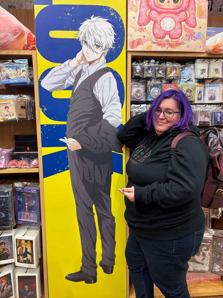 Image of TJ, a light-skinned nonbinary person with purple hair, posing in front of a poster of Nagi Seishiro from Blue Lock.