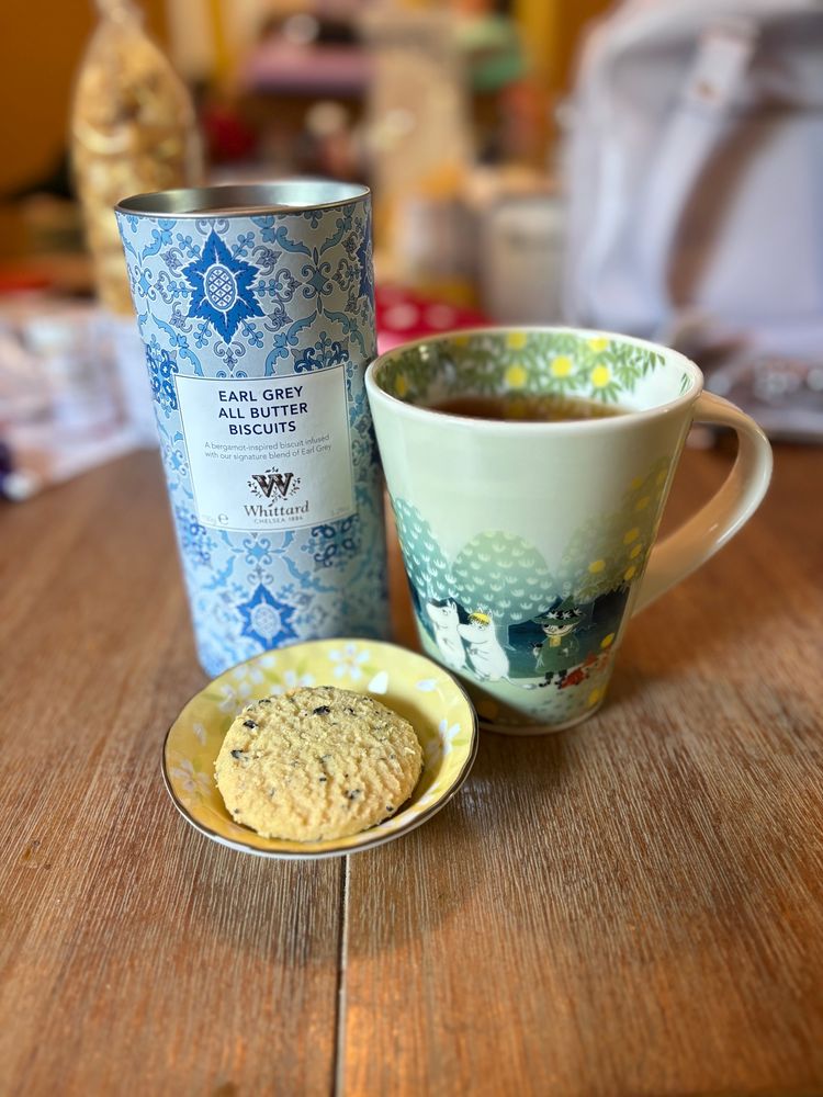 Photo of TJ’s Moomin mug, featuring Moomin, Snorkmaiden, and Snufkin in the Moominvalley forest, filled with tea next to a small round, yellow dish of butter cookies and a blue tin labeled “Earl Grey All Butter Biscuits.”