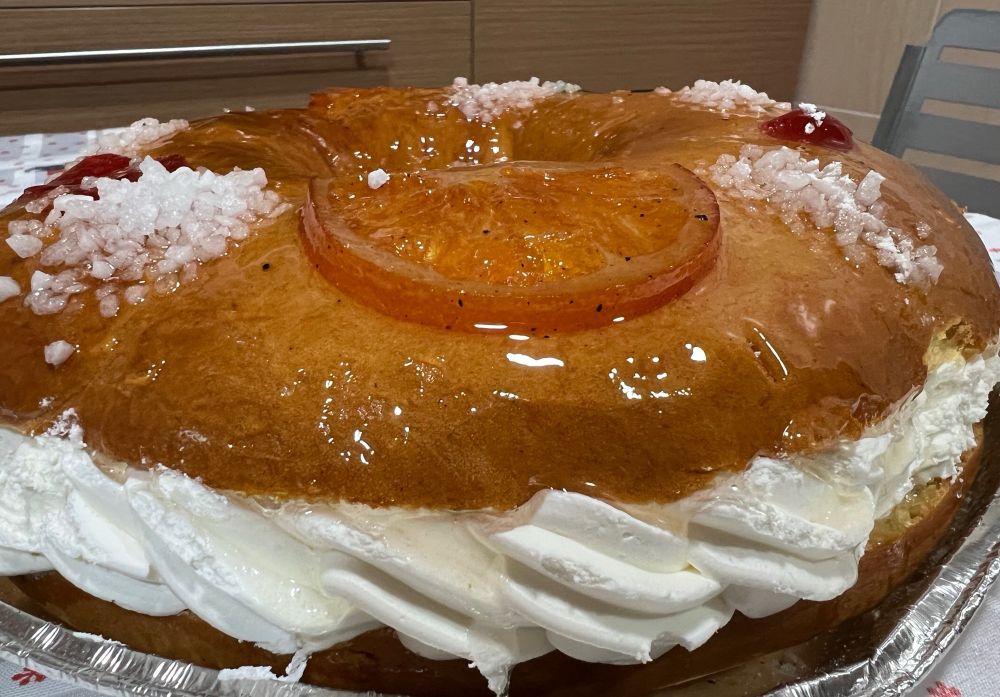 Un Rosco de Reyes relleno de nata. Vista lateral, se ve el azúcar, brillante y también se aprecia un trozo de naranja confitada de adorno. 