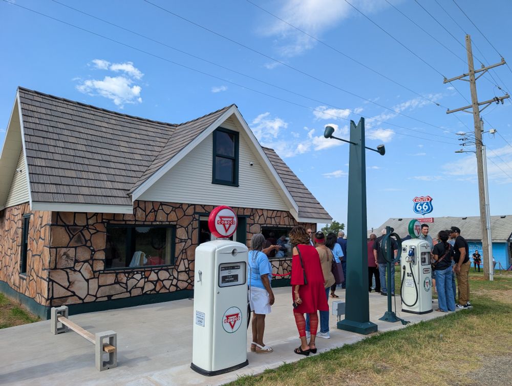 A stone gas station with vintage gas pumps out front. A large gathering of people surrounds the station.