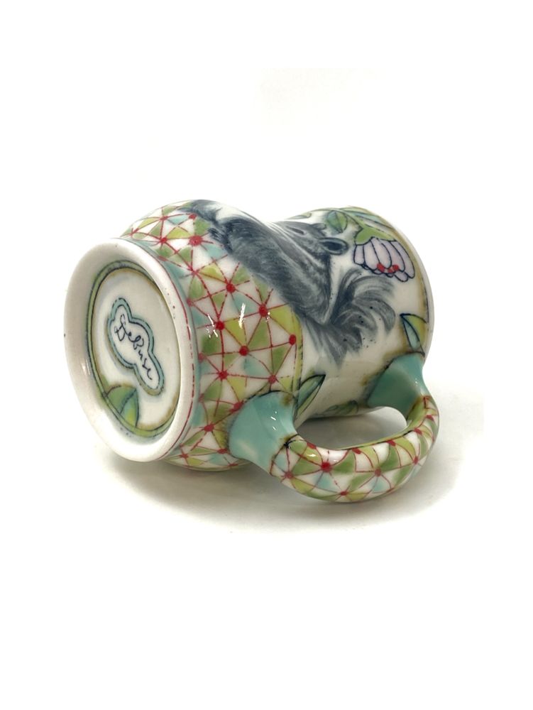 A handmade mug on its side where the handle and base are visible, revealing a geometric pattern in greens with red dots.
