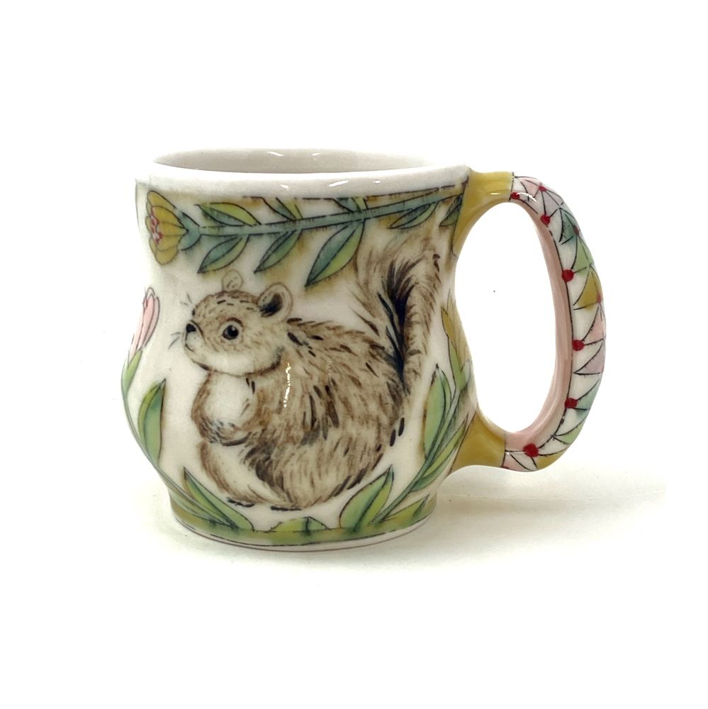 A handmade porcelain mug with a painting of a brown chonky squirrel flanked by flowers in greens and yellows.