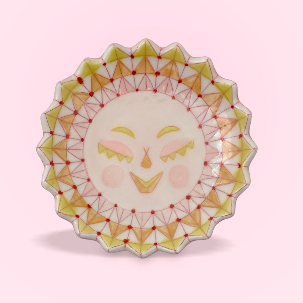 A small ceramic plate decorated like a stylized sun on a pink background.