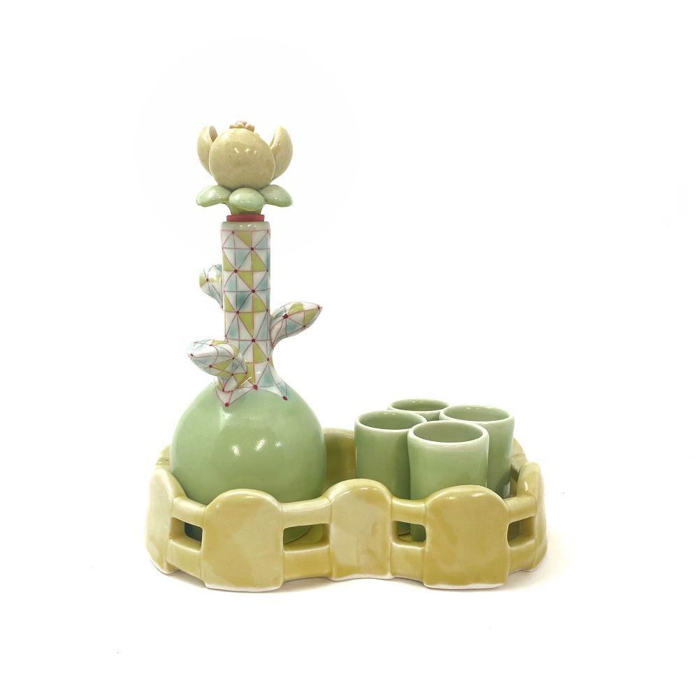 This liquor set features a bottle that resembles a flower. It has a green bulbous base, a tapered neck with a geometric pattern and the bottle stopper is flower-shaped with green and yellow petals. The set features four green sipper cups. The bottle and four cups sit in a yellow fence tray.