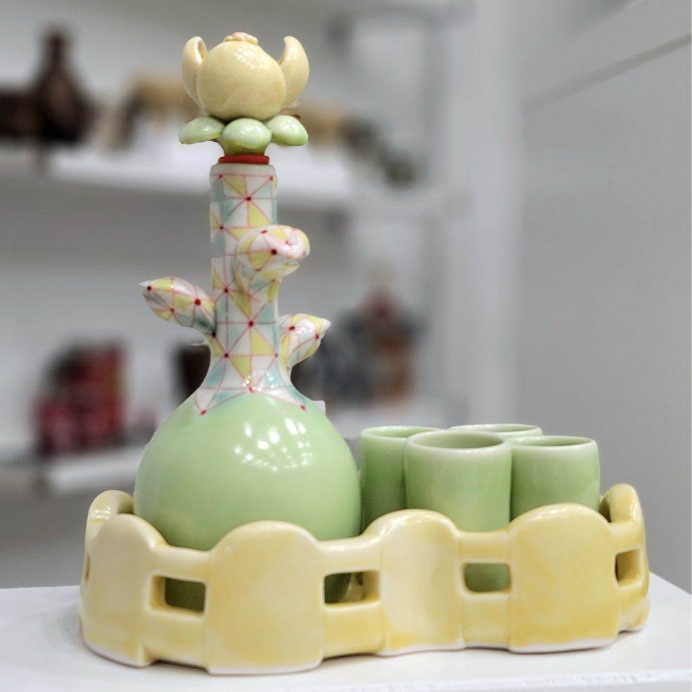 A liquor set with a bottle shaped like a stylized flower and four small green cups sitting in a yellow fence tray. 
