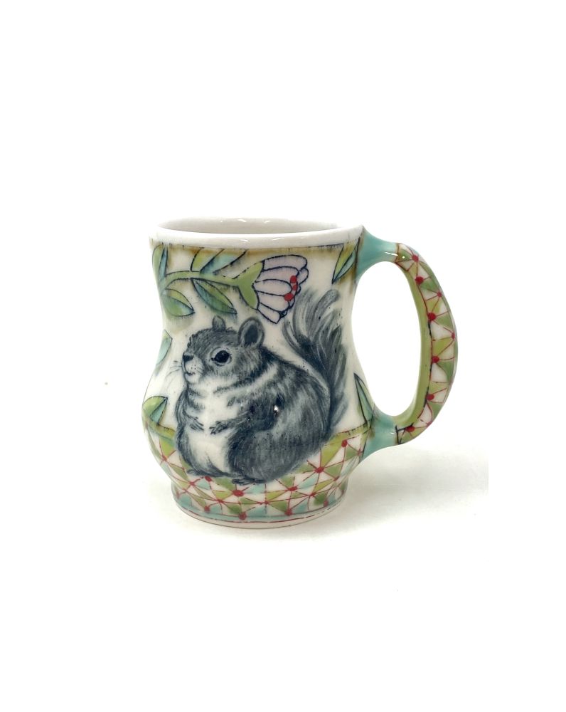A handmade porcelain mug with a painting of a chonky squirrel flanked by flowers and a geometric pattern in greens, pink and red.
