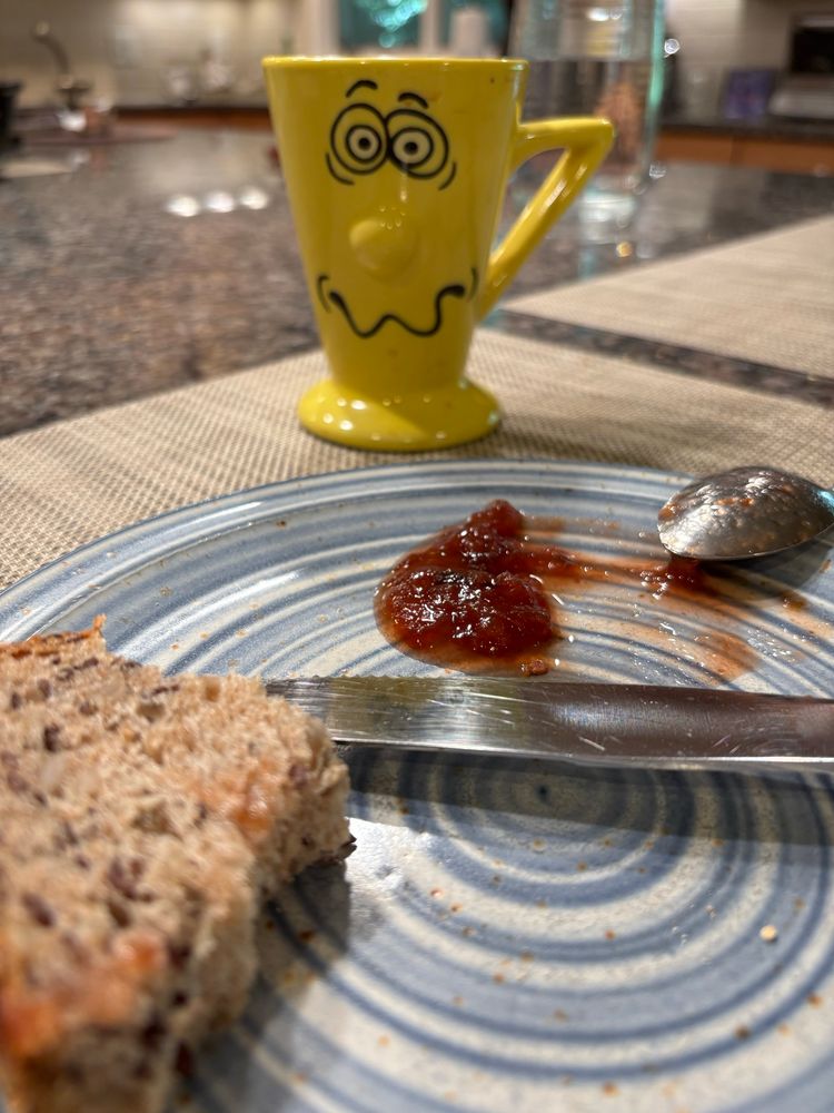 A yellow cup (of coffee) with a dazed, stressed & confused face drawn on it in black. This guy needs help! In the foreground a half-eaten piece of bread & some plum jam on a blue & white plate.