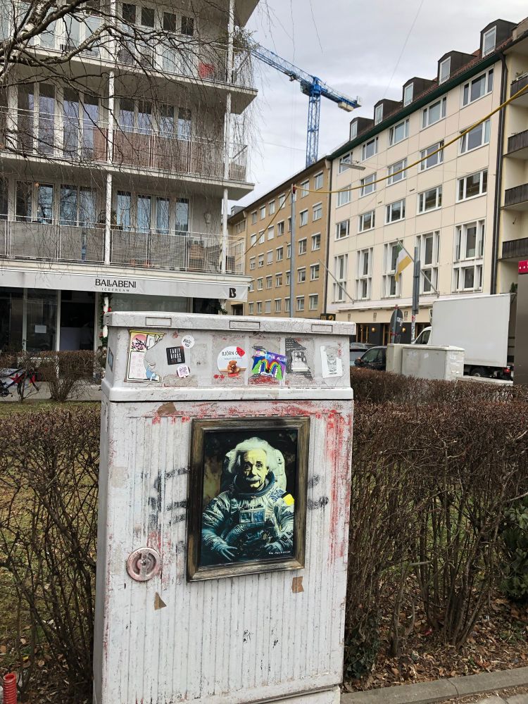A very random city snapshot taken in Munich; in the foreground a transformer (electrical box) at the edge of a sidewalk w/stickers, spray-paint & a framed portrait of Einstein in a spacesuit; behind Apollo 13-Albert there are apartment buildings, a glimpse of a crane, & the Ballabeni ice cream store w/out its usual long queue (because it’s closed in winter). 