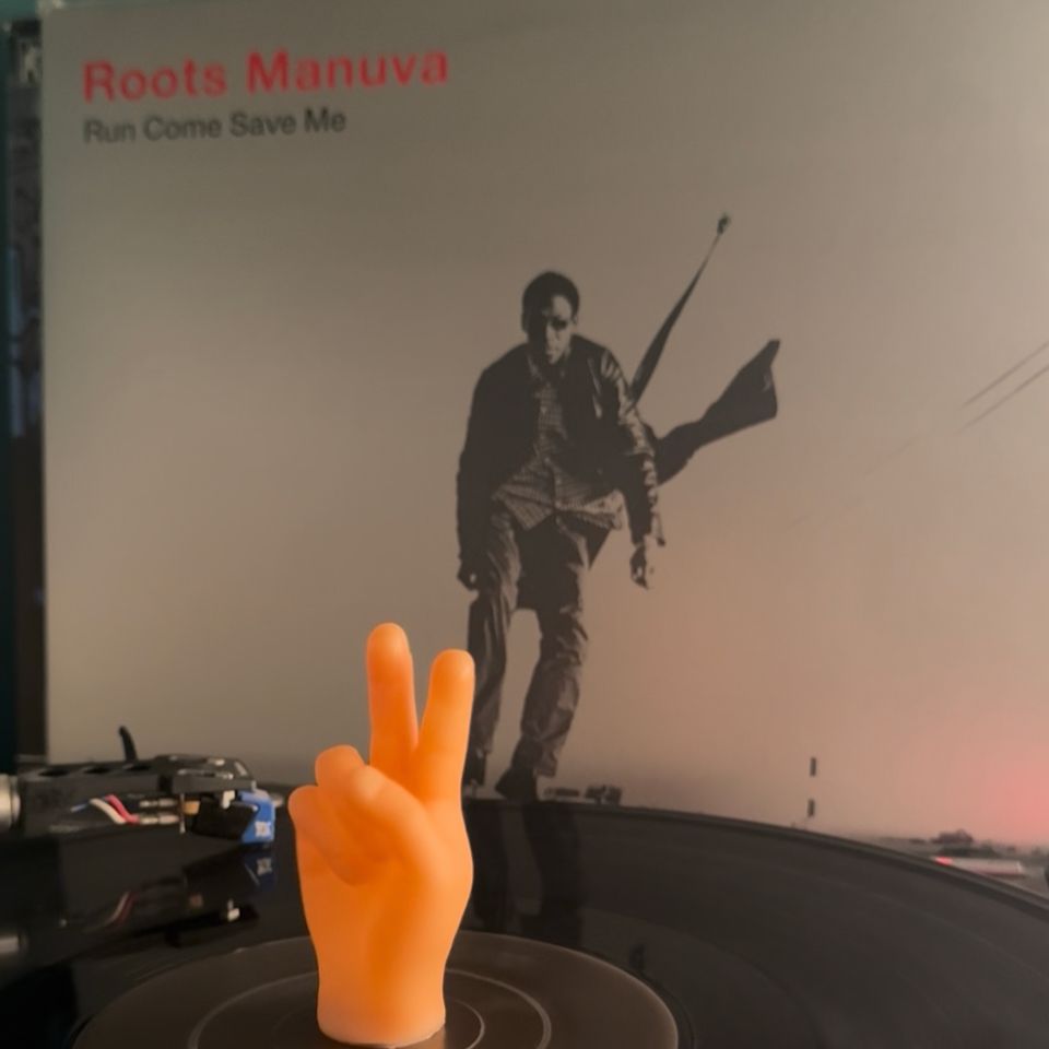Picture of the Roots Manuva- Run come save me, album cover. Black and white photo of the man himself jumping in the air on a deserted road. The vinyl is in the foreground on a turntable, a tiny hand doing a peace sign is spinning in the centre. 