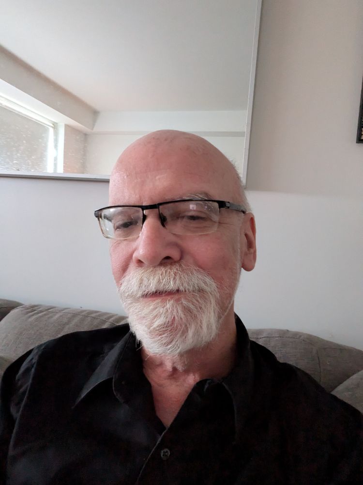 Bald white man with glasses, a white beard around his upper and lower lips, and a black shirt.