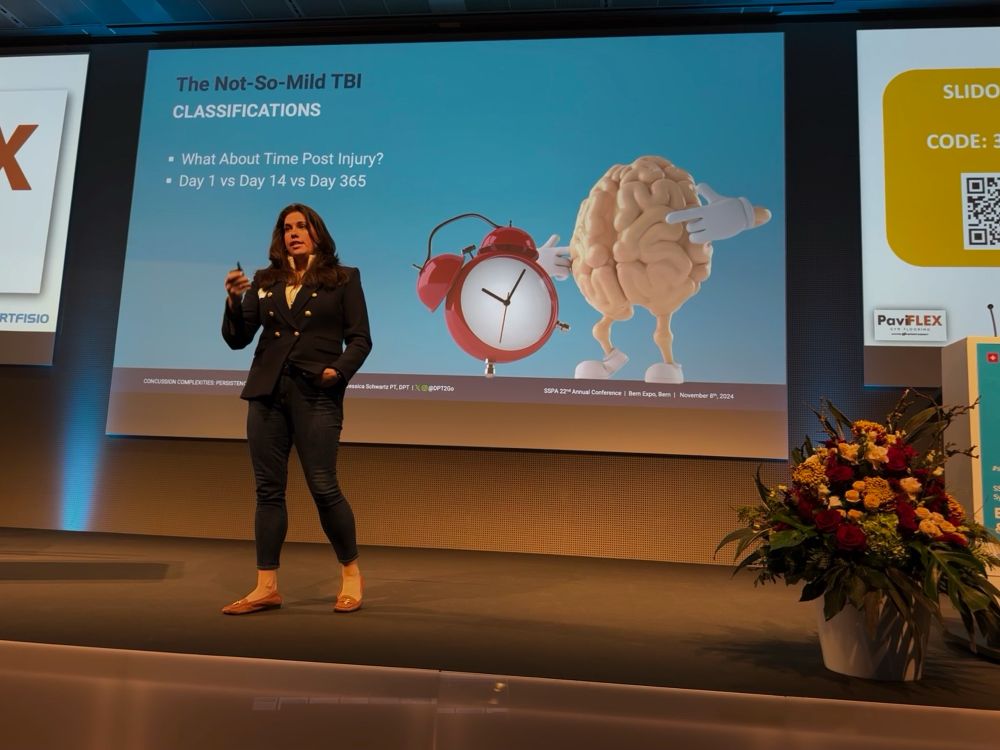 Jessica Schwartz, Physical Therapist and Brain Injury Survivor, delivering a lecture in a blue suit to 500+ Fisio’s on Concussion Complexities with a blue slide behind her discussing the importance of time post injury 