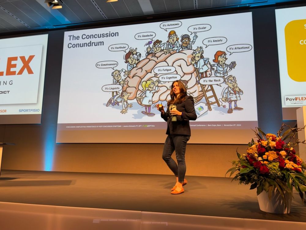 Jessica Schwartz, Physical Therapist and Brain Injury Survivor, delivering a lecture in a blue suit to 500+ Fisio’s on Concussion Complexities with her concussion cartoon she developed in 2015 behind her on a slide