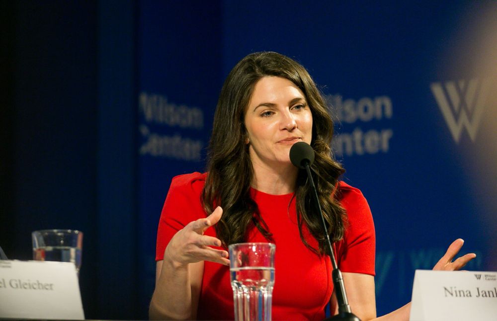 Nina Jankowicz in a red dress speaking on a Wilson Center panel in front of a blue backgriund with the wilson center logo on it 