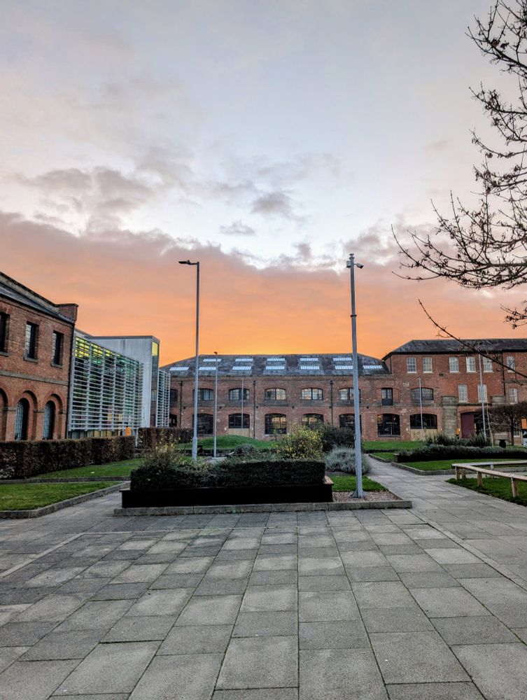 A red sunrise above a Victorian railway building 
Modern paving slabs in the foreground and shrub beds in the middle 