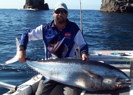 An angler posing with a recently caught Gasterochisma specimen aboard a small fishing vessel close to some rocks. The scales are so shiny you can see reflections in them. The fish is quite big, perhaps 150cm long, and apparently weighed 32kg. Credit: Wade Pelham, RedMap.