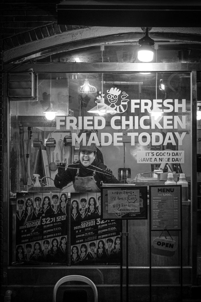 A black and white photo taken in Seoul. A Korean woman in the front window of a fried chicken shop smiles and poses playfully with a set of tongs in each hand. The window has lettering applied that reads ‘Fresh Fried Chicken Made Today’ and in smaller type beneath: ‘It’s a good day. Have a nice day.’
