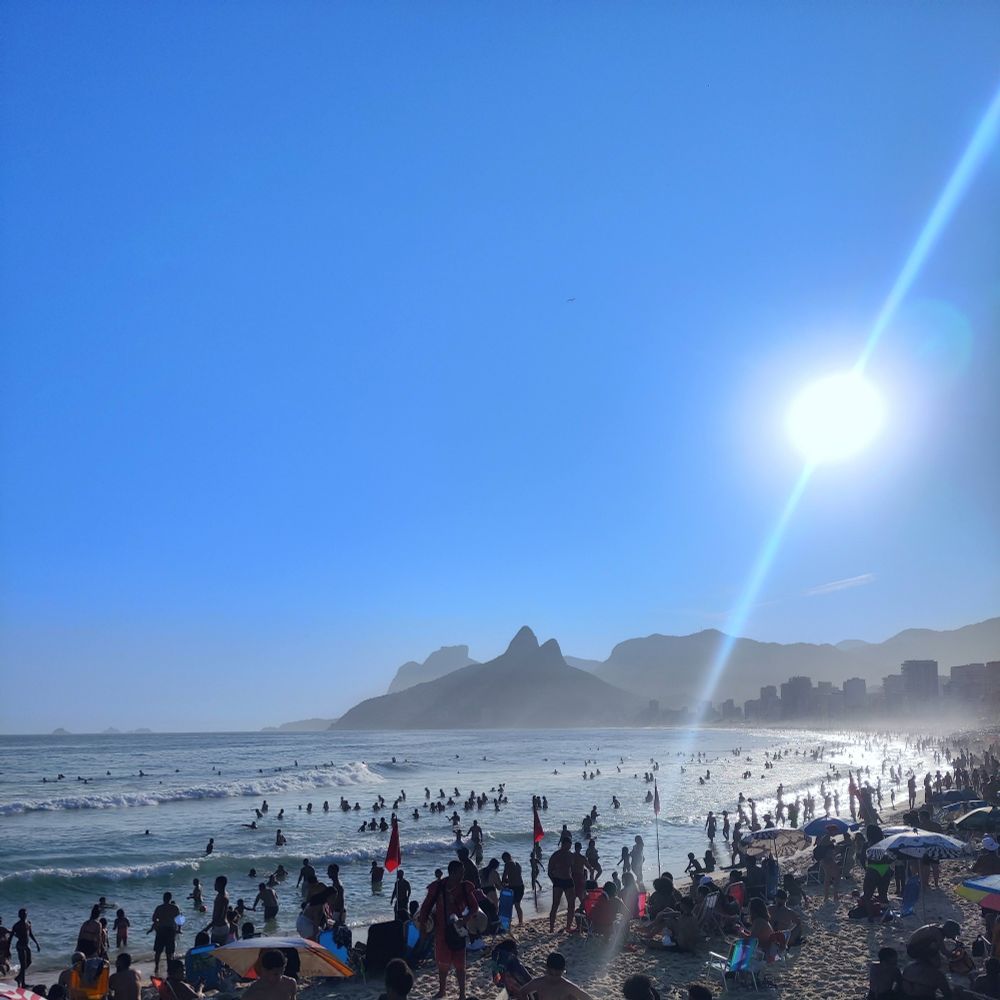 Imagem do Rio de janeiro, focada em mostrar o céu azul, pessoas na praia e montanhas ao fundo