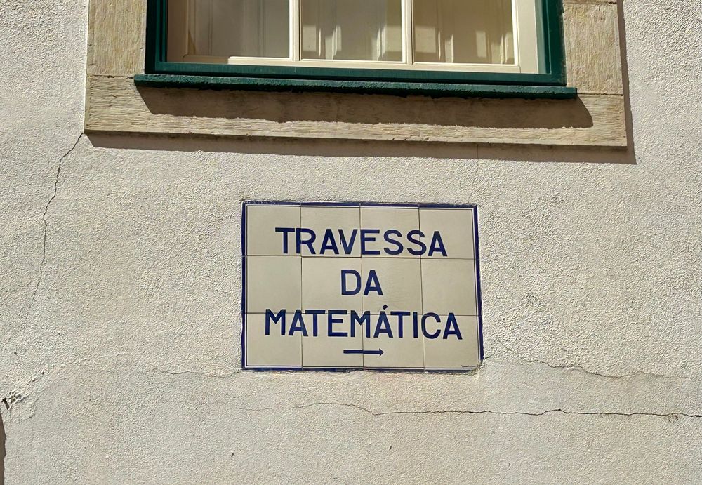 A blue and white tiled street sign on a white wall. It reads “Travessa da matemática” which translates to something like “cross street of mathematics”.