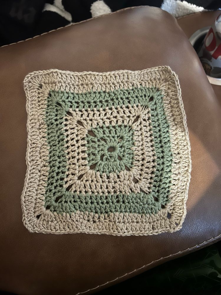 A crocheted granny square in a solid granny square pattern. The middle is sage green, then alternates colors every two rows between beige and sage green. 