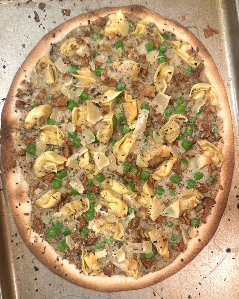A vegan pizza served on a metal baking sheet, featuring a homemade white sauce base topped with crispy fried tortellini, crumbled tempeh bacon, vibrant green peas, and a sprinkle of herbs. The crust is golden brown and slightly charred at the edges.