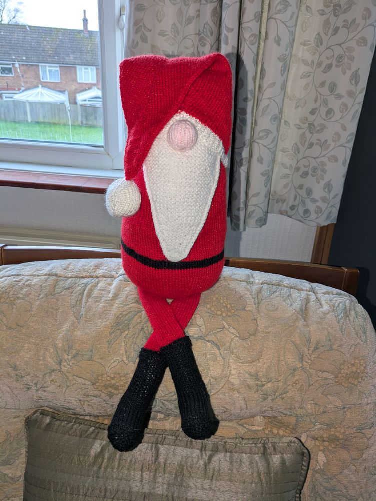 All three shots show a gonk style santa. He has a droopy hat and long flat legs.
In this picture he is perched on the back of an armchair with his legs crossed. 