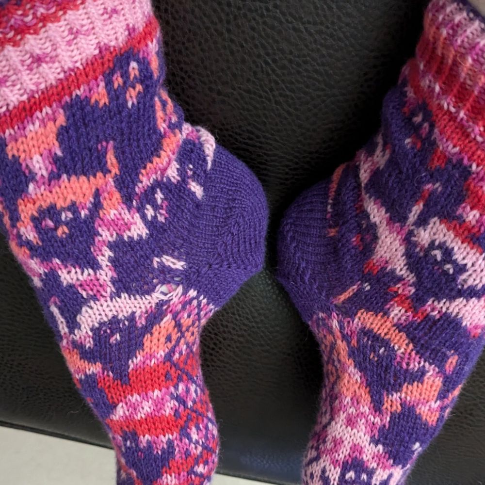 A pair of handknit colourwork socks.
Purple ghosts with a pink self striping background.
This view is a side view. You can see a purple shirt row heel and a criss cross pattern on the sole. 