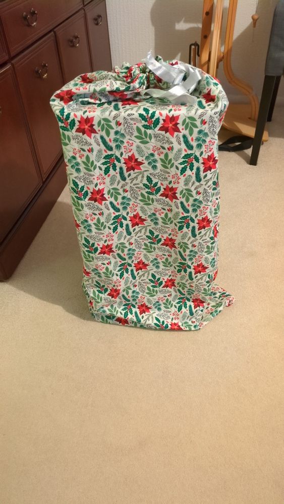 A large fabric drawstring bag which has been made with Christmas fabric (poinsettias and other foliage)
There is a present in the bag but you can't see what it is.
There is a spinning wheel in the background, the present is almost as tall as the spinning wheel. 