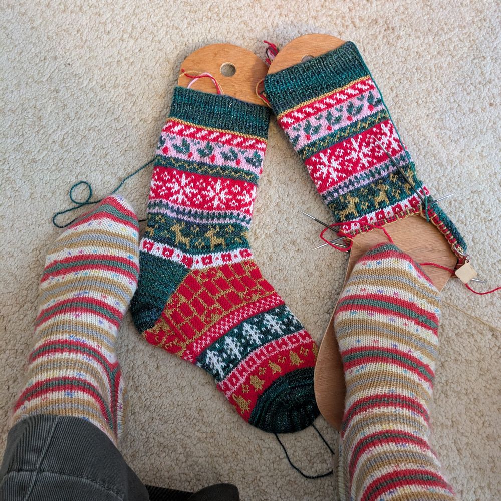 A pair of feet wearing handknit self striping Christmas socks with a pair of half knit colourwork Christmas socks on sock blockers in the background. 