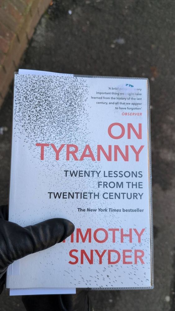 The book On Tyranny by Timothy Snyder is held in the leather gloves hand against a pavement background