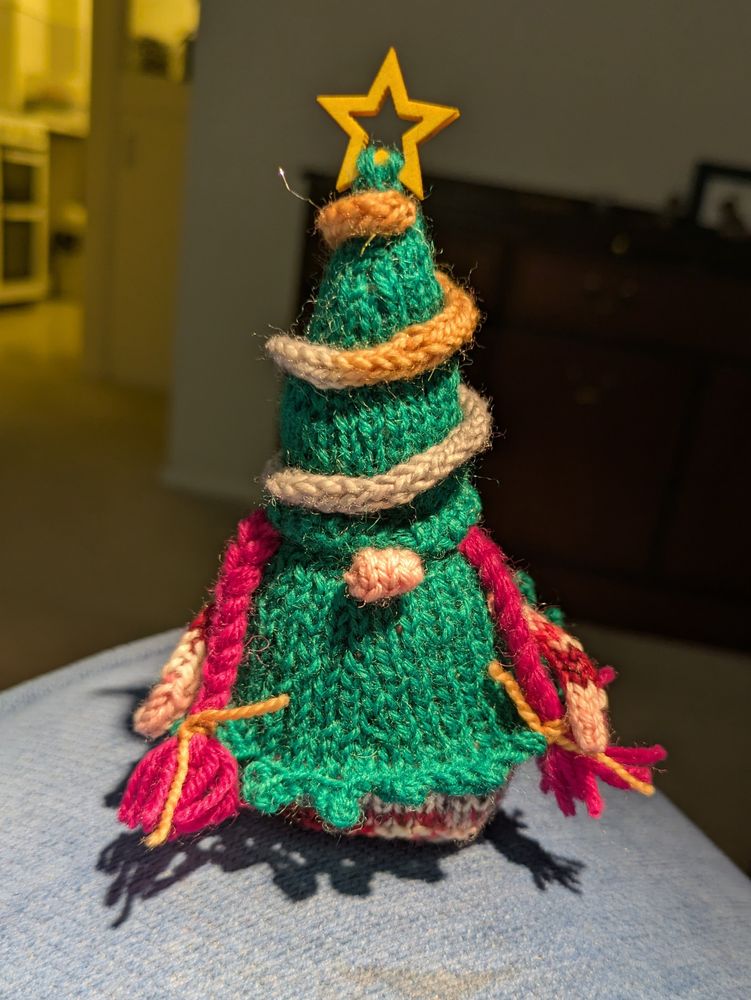 A hand knitted gnome dressed as a Christmas tree, with a green hat and dress. Tinsel around the hat and a star on the top.
She has bright pink braids. 
