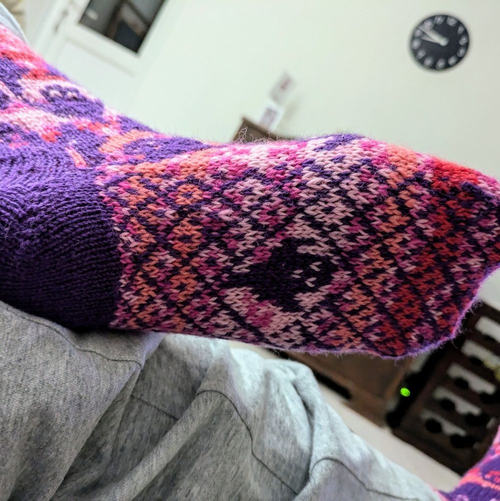 A pair of handknit colourwork socks.
Purple ghosts with a pink self striping background.
This view is of the sole of a single sock. It has a criss cross pattern a small ghost motif in the centre of the sole. 