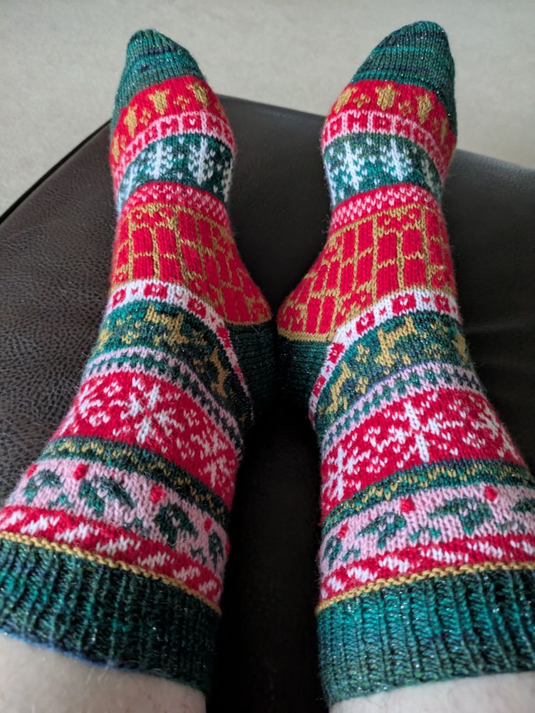 A pair of multi motif colourwork Noridc style handknit socks in dsrk green, pink, red, gold and white.
Motifs include holly, snowflakes, reindeer, presents, trees, Ho ho ho and bells. 