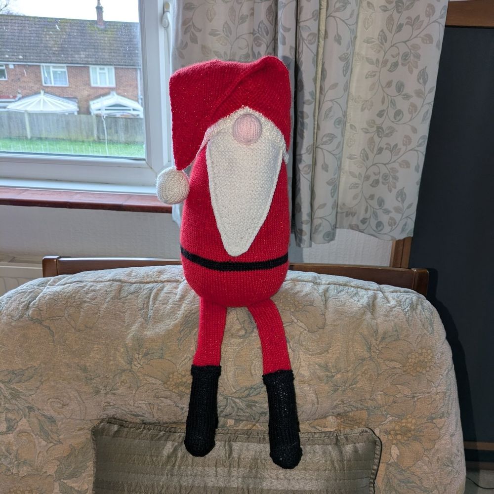 All three shots show a gonk style santa. He has a droopy hat and long flat legs.
In this picture he is perched on the back of an armchair with his legs dangling straight. 