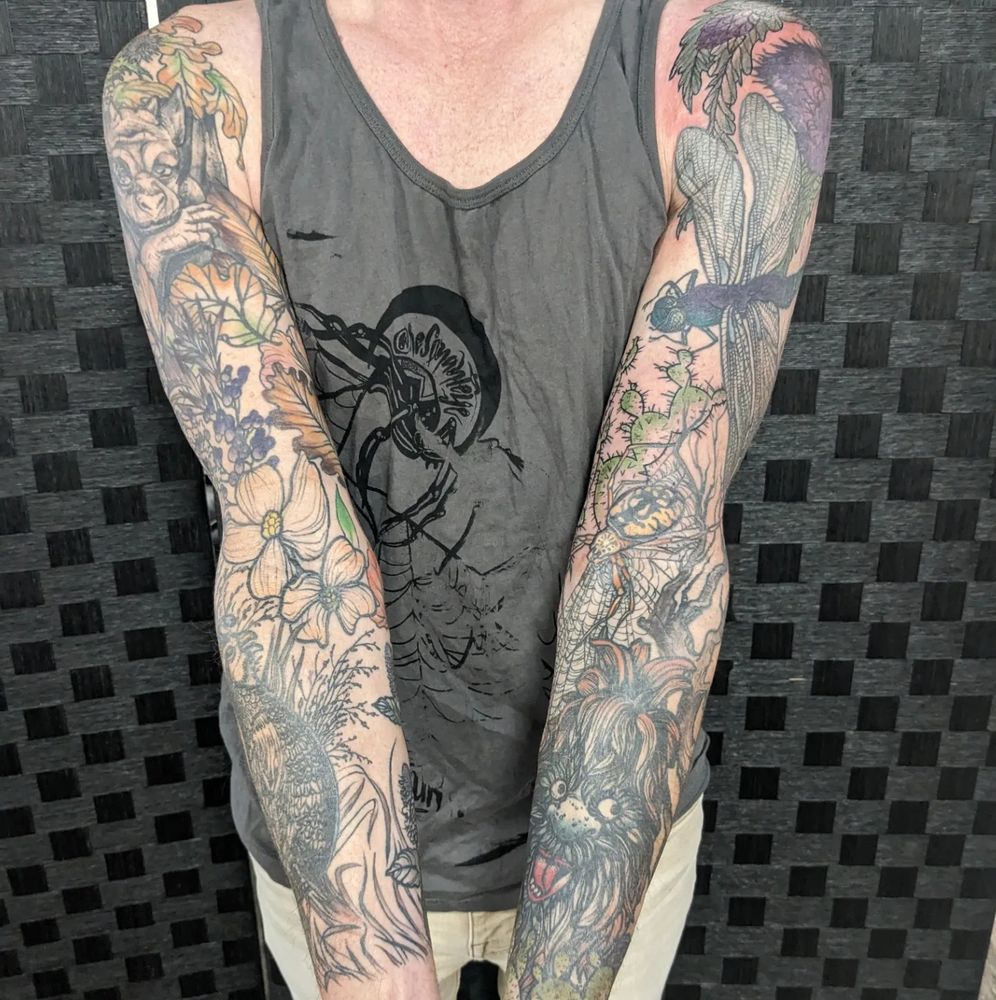 someone in a tank top with their arms extended to show full sleeve tattoos. there are flowers, dragonflies and a silly creature