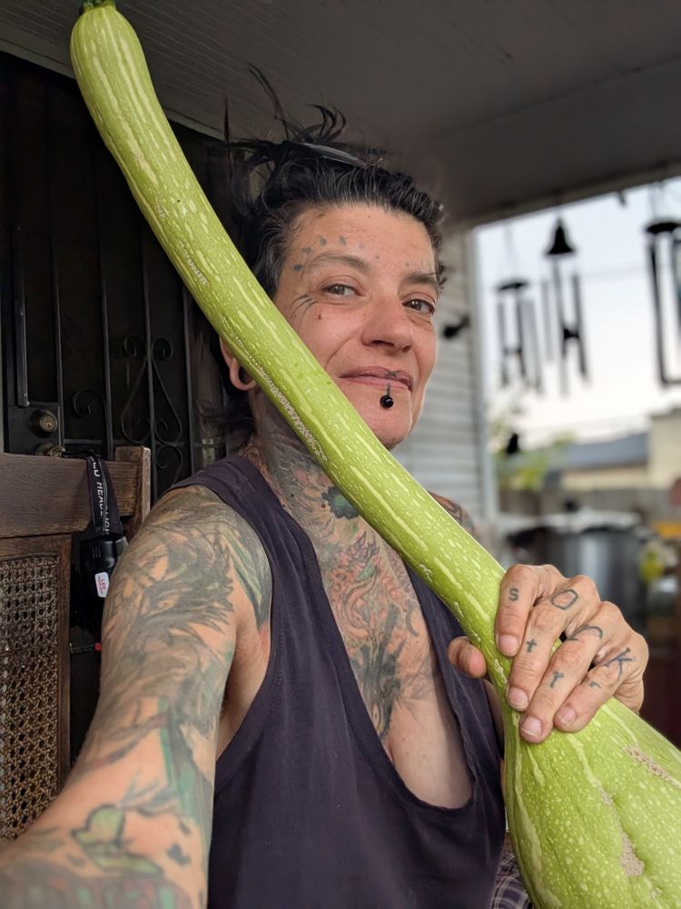 a tattooed person holds a large tromboncino squash towards the camera