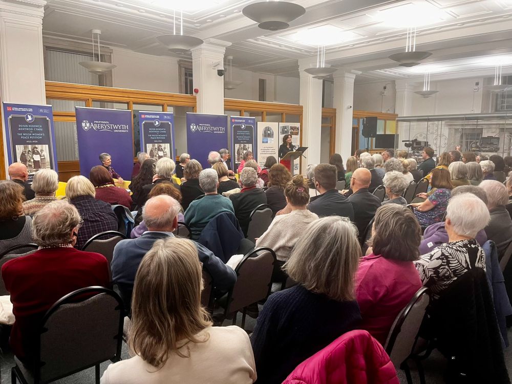 Photograph of a large room, filled with an audience in rows of chairs watching a woman speak at a lectern in front of banners about the Welsh women's peace petition of 1923-24