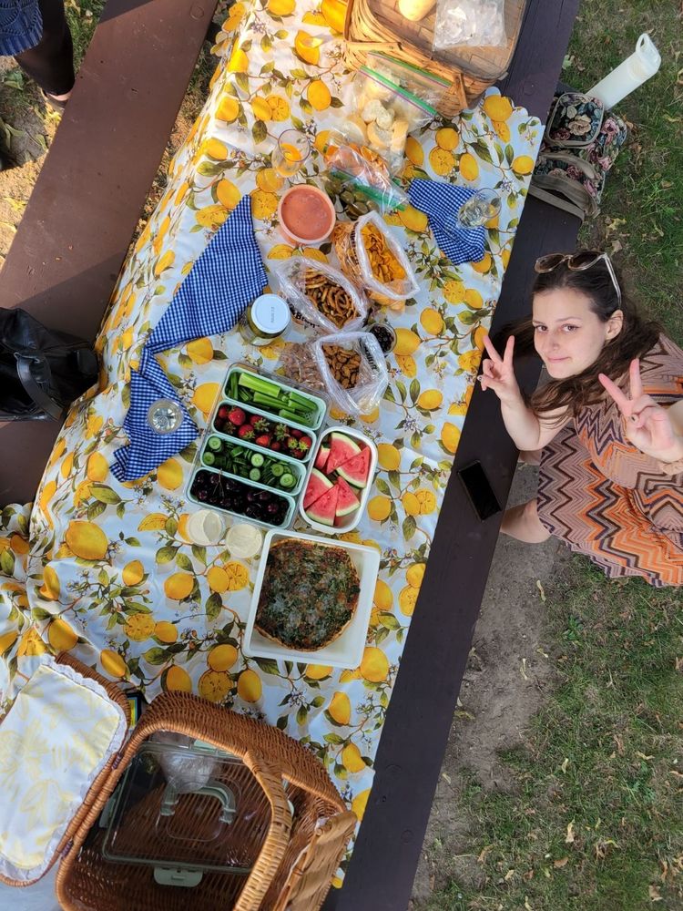 picnic spread on picnic table, taken at a top-down angle by a friend standing on the bench. I am standing next to the table, bending over to get in frame, looking up at the camera and making a peace sign with both hands. 
the picnic consists of a spinach pizza, hummus, cucumbers, celery, cherries, strawberries, watermelon, dill pickles, garlic sesame sticks, pretzels, jalapeno cassava chips, roasted tomato and garlic soup, feta stuffed olives, oranges, homemade bread, dark chocolate covered coffee beans, and white wine.