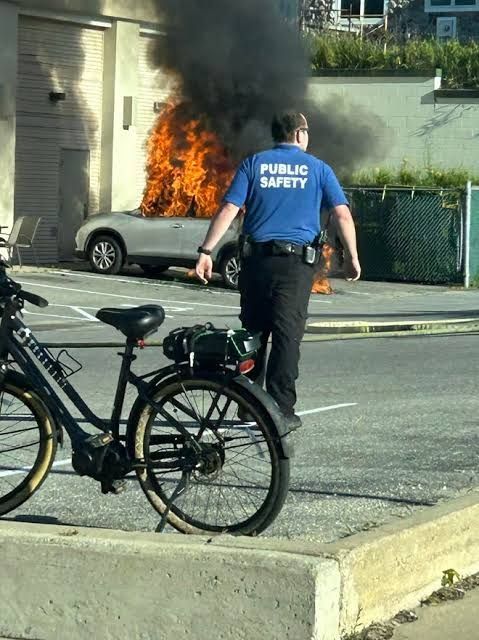 A man with a tee shirt emblazoned "public safety" walks from his bicycle towards a burning car.