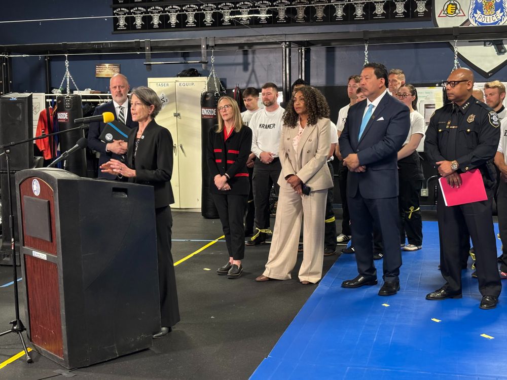 Council President Nelson at a podium speaking. Behind her are Councilmember Bob Kettle, Erin Goodman, Executive Director SODO BIA, Monica Alexander, Executive Director WSCJTC, Seattle Mayor Bruce Harrell, Seattle Police Chief Shon Barnes, and many new police officers.