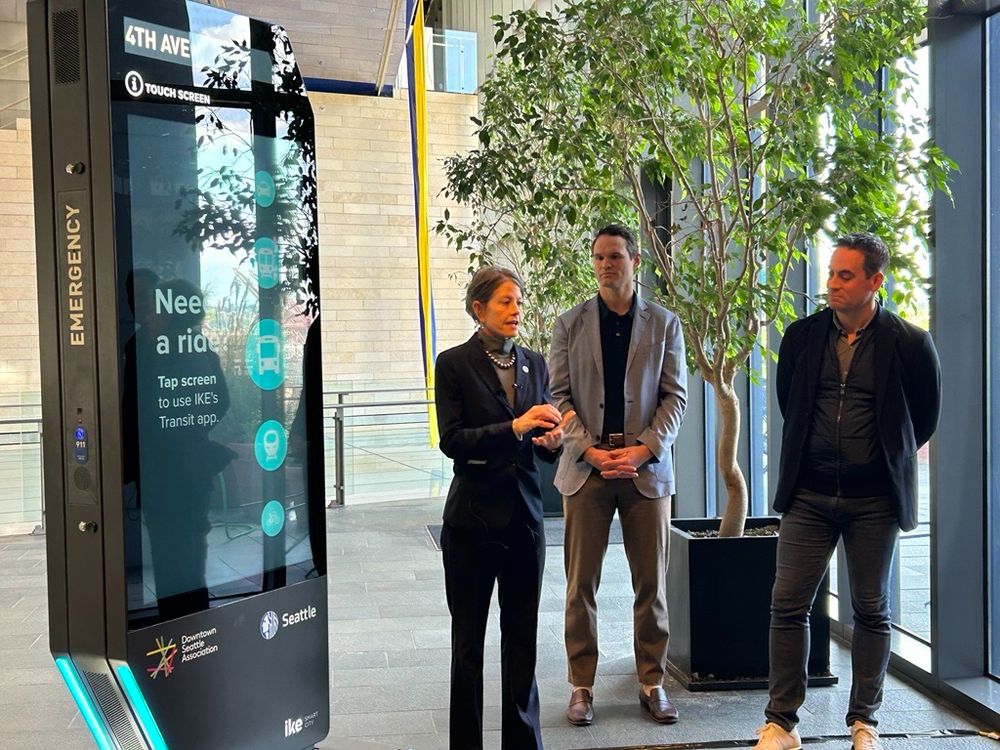 Council President Sara Nelson Speaking next to a digital kiosk showing bus information on the screen. 