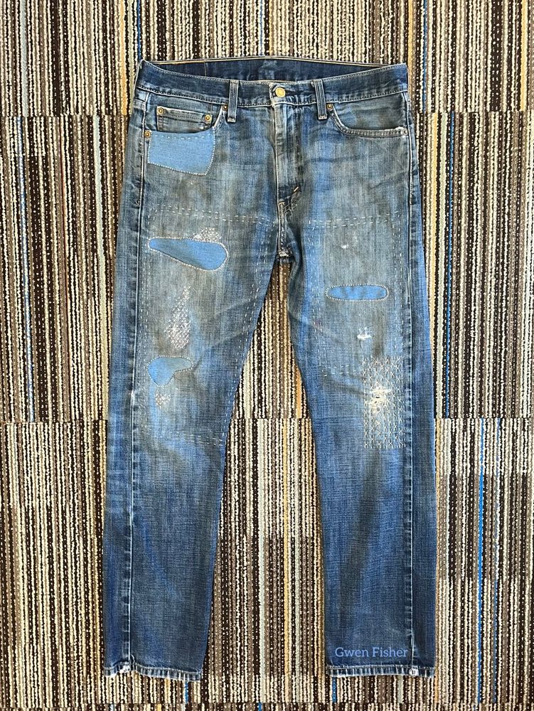 Photo of old jeans lying flat on carpet. The jeans are mended with various techniques including inside patching, outside patching and sashiko. 