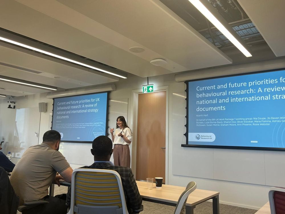 An image of a woman standing at the front of a room, in front of two projections where the text says 'Current and Future Priorities for UK Behavioural Research. A review of national and international strategy documents.'