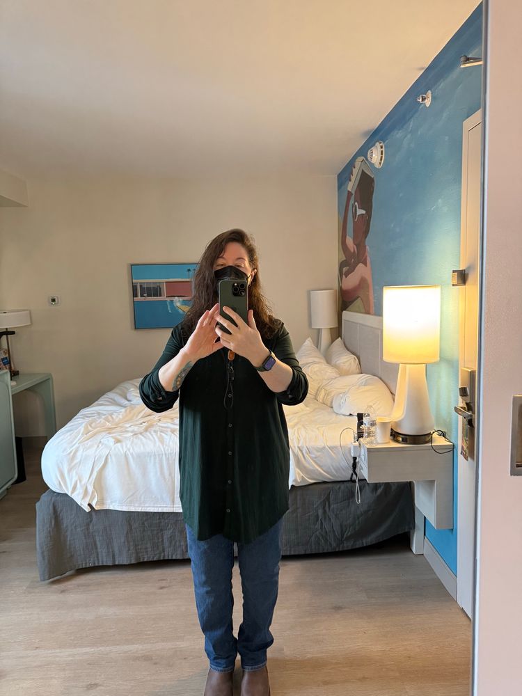 A while woman in jeans and a dark top, with damp hair and a black ffp3 mask, takes a selfie in front of an unmade bed in a very Florida looking hotel room 