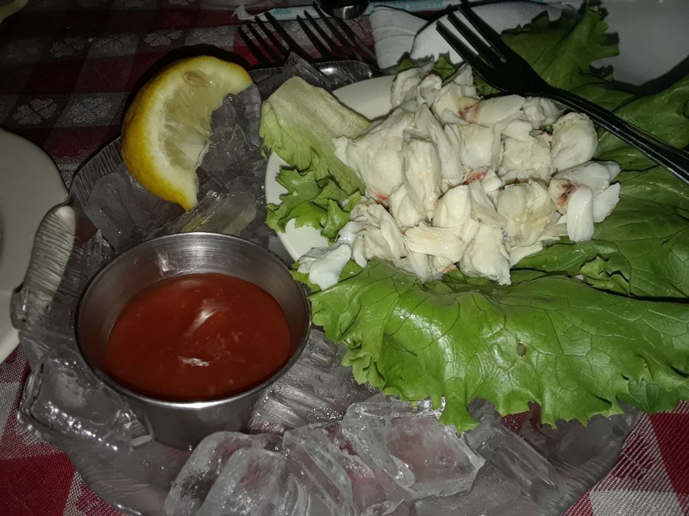 Picture of crabmeat cocktail, with crabmeat and lettuce in the right third, cocktail sauce in the lower left third, and a lemon in the upper right third, all surrounded by ice and some forks at the top.