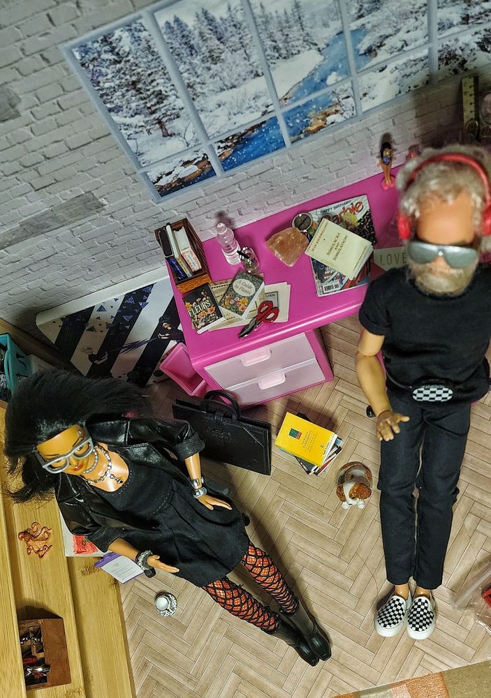 Two dolls pursuing blissed together. In this picture, shot from above, they are in an office space, with a snowy view out the window. 