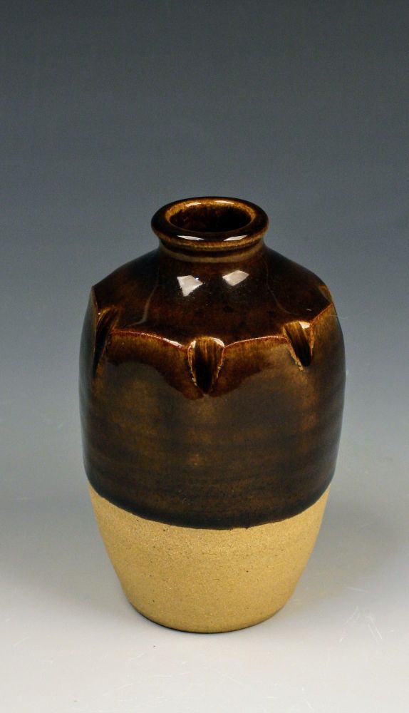 Small ceramic bottle with brown glaze. Divots tapped into the top to create interest in seven sided fashion.